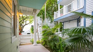 A covered wooden deck with a white railing, surrounded by lush green plants and a white picket fence. - Ecco Domani by AvantStay | Gorgeous Home w/ Pool, Balcony in Great Location (Key West)