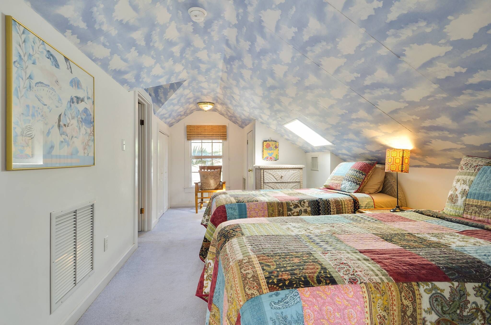 Un dormitorio con techo inclinado, una cama grande con colcha colorida, una mesita de noche con lámpara y una ventana con persianas.