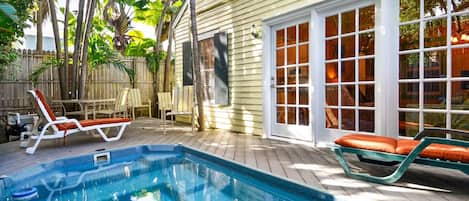 Une piscine avec deux chaises longues, une terrasse en bois et une maison dotée d’une grande fenêtre et d’une porte.