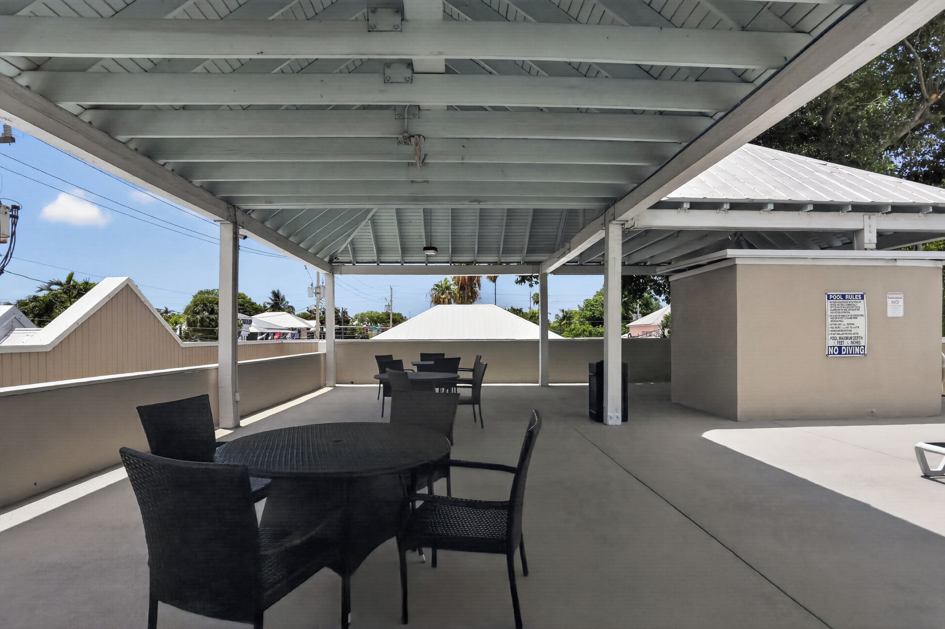 An outdoor covered patio with tables and chairs, a white building in the background, and a sign indicating pool rules.