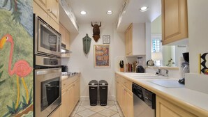 A kitchen with wooden cabinets, a microwave, a stove, a sink, and two trash bins. - Truman's Hideaway | Top Location w/ Patio, Pool Access & BBQ | Month Long Stays (Key West)