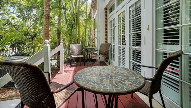 A covered outdoor area with wicker furniture and a mosaic table, surrounded by lush greenery and a white building with large windows. - Pool + Walk to Duval & Fort Zachary | Monthly Stays | Key West Found (Key West)