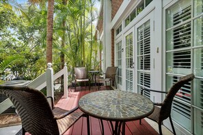 Un espace extérieur abrité, aménagé avec des meubles en osier et une table en mosaïque, entouré d’une végétation luxuriante et d’un bâtiment blanc doté de grandes fenêtres.