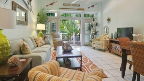 A spacious living room with a sofa, armchairs, a coffee table, and a television. There are decorative items like a fish sculpture and a mirror. The room has a ceiling fan and a view of the outdoors through large windows. - Pool + Walk to Duval & Fort Zachary | Monthly Stays | Key West Found (Key West)