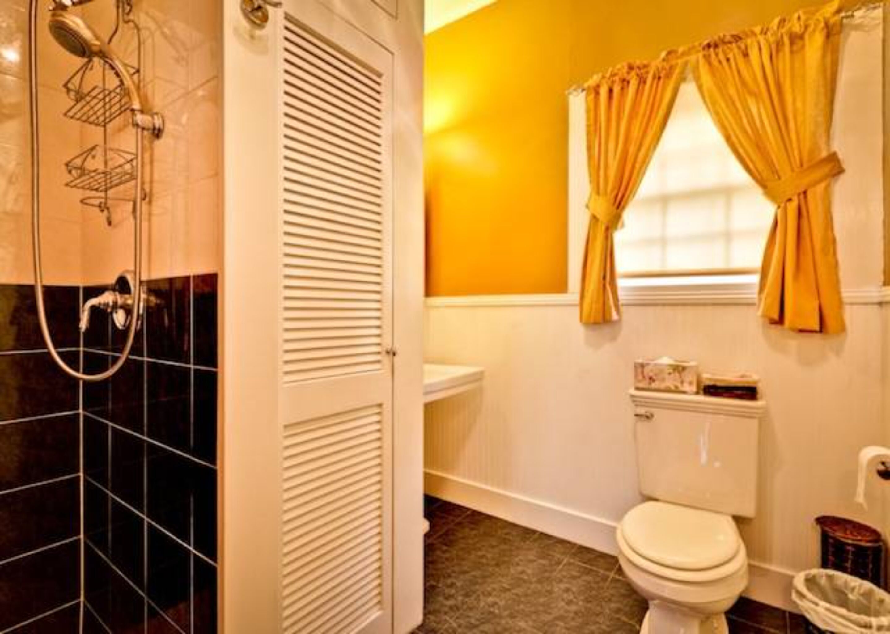 A bathroom with a shower, toilet, and a window with yellow curtains.