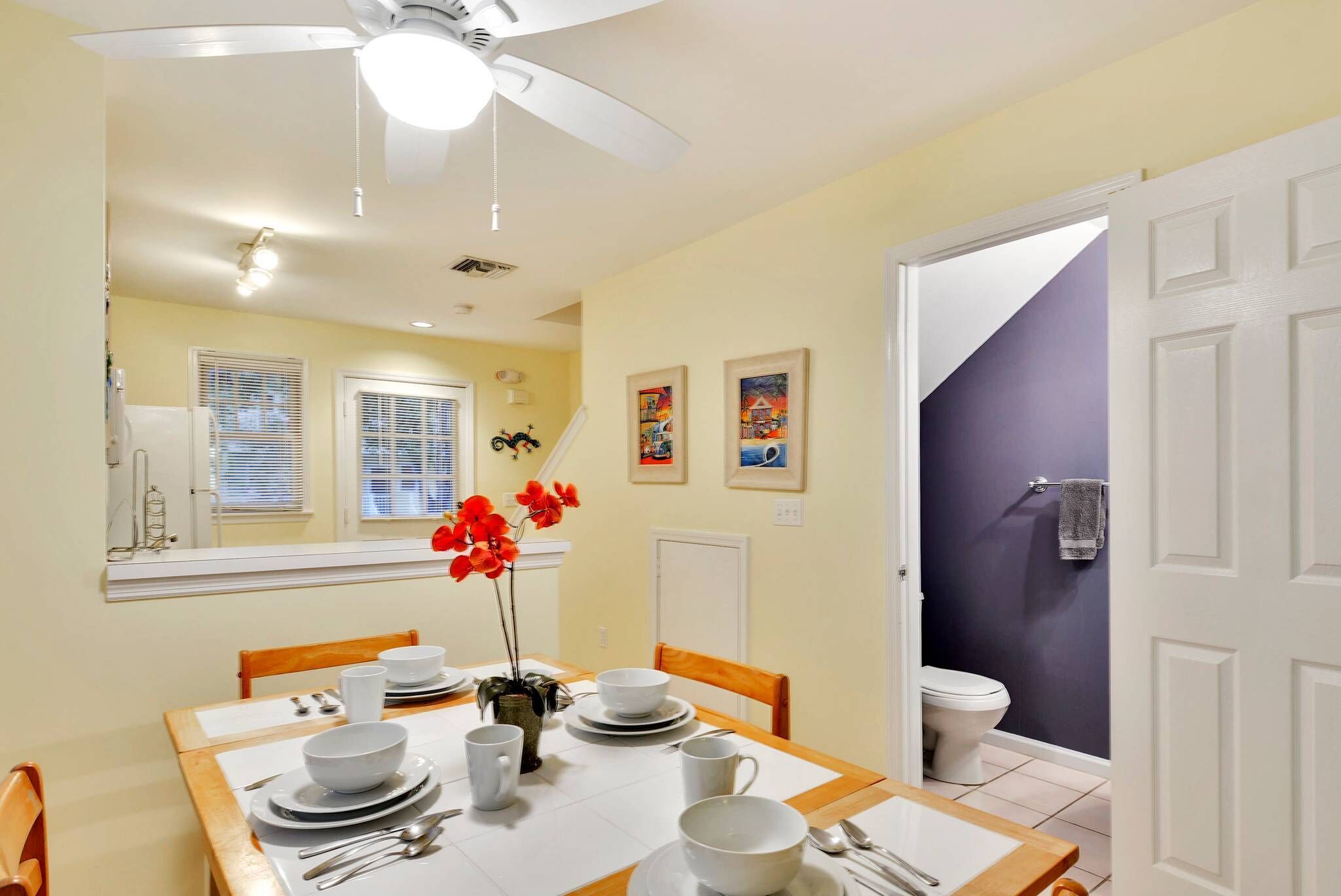 A dining area with a table set for four, a ceiling fan, and a view of the bathroom.