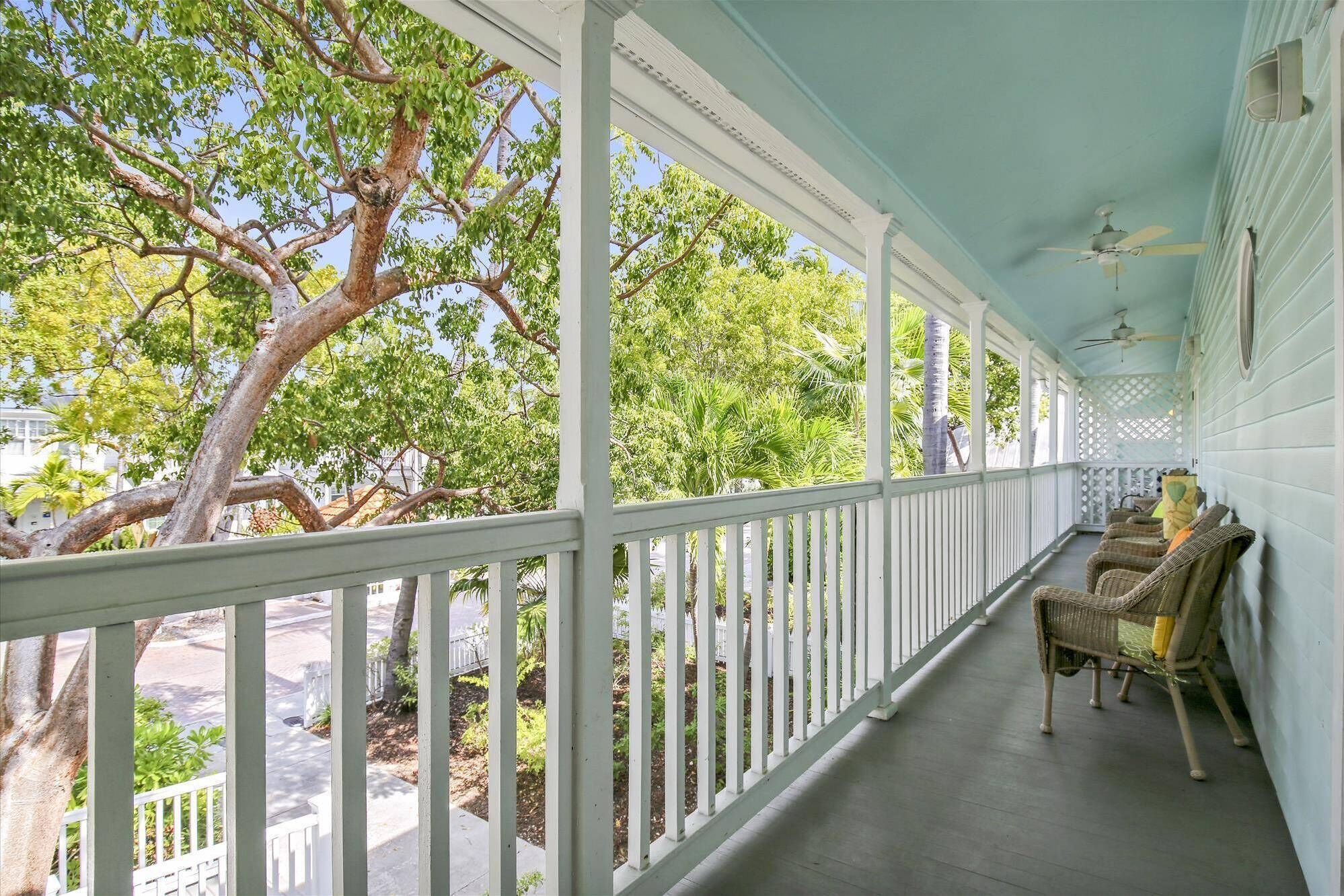 A covered porch with white railings, wicker furniture, and a view of trees and a street.