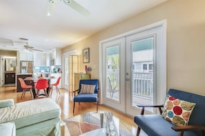 A spacious living room with a large window, a dining area with red chairs, and a kitchen in the background.