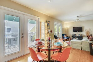 A dining area with a glass table and orange chairs, a living room with a sofa and TV, and a balcony with blinds.