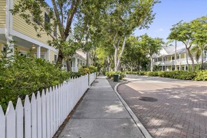 A tree-lined street with a white picket fence, a sidewalk, and a brick-paved road.