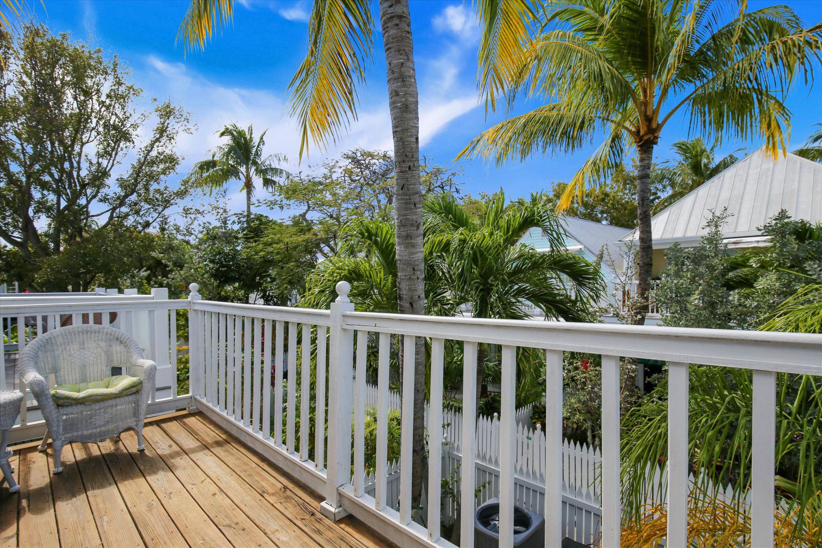 A wooden deck with white railings, a wicker chair with a cushion, and a view of palm trees and a house in the background.