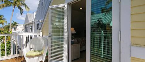 A wooden deck with a white wicker chair, a sliding glass door leading to a bedroom, and a palm tree in the background.