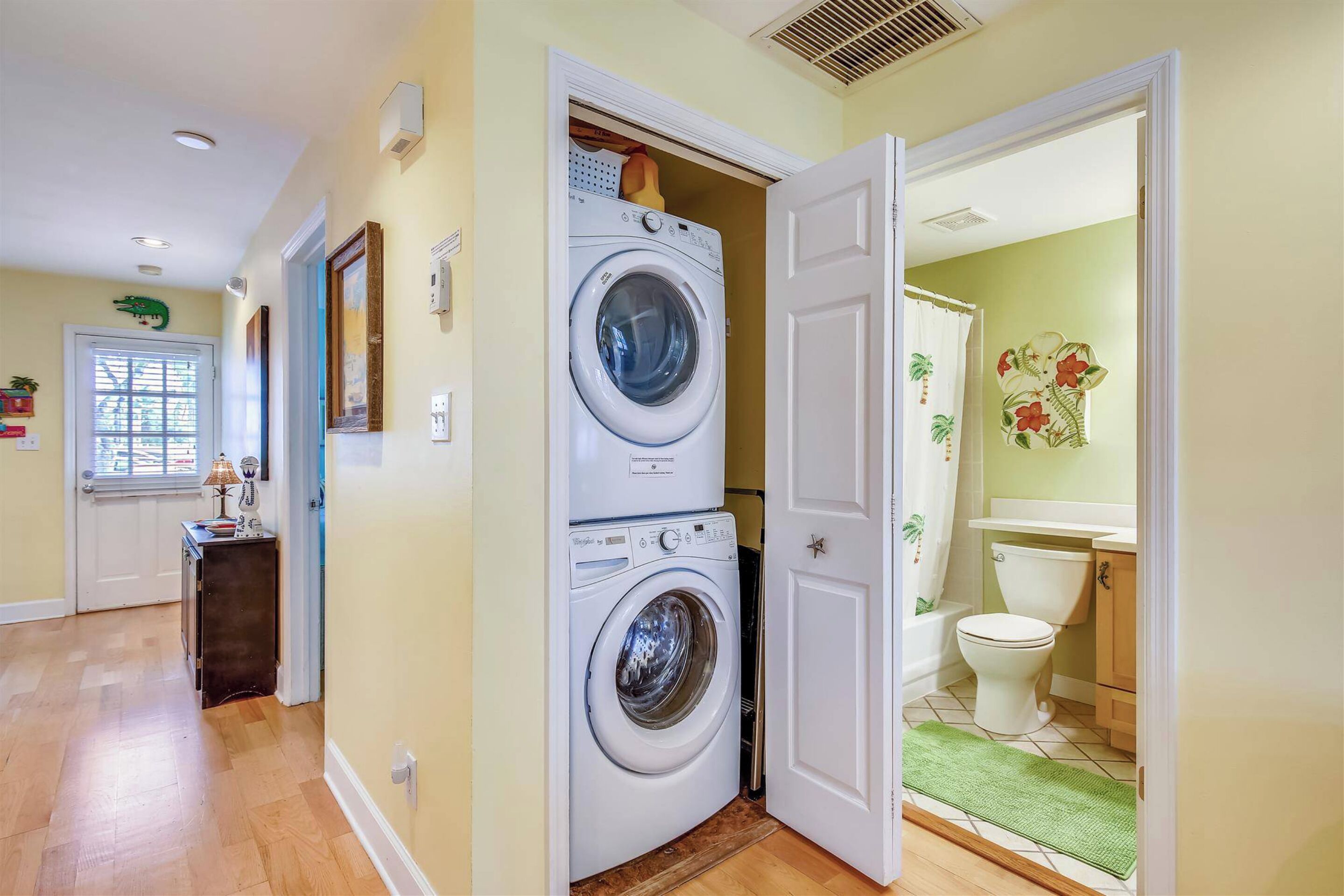 Une buanderie équipée d’un lave-linge et d’un sèche-linge superposés, une porte donnant sur une salle de bain avec des toilettes et un lavabo, et un couloir avec une lampe et une petite table.