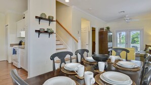 Un comedor con una mesa de madera dispuesta para cuatro personas, rodeada de sillas. Se ve una escalera en el fondo que conduce a otra habitación.