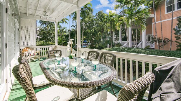 A covered outdoor area with a glass table and wicker chairs, surrounded by a white railing, palm trees, and a building in the background.
