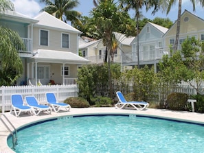 A swimming pool with lounge chairs, surrounded by a white picket fence, with houses in the background.