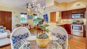 Cozinha moderna com eletrodomésticos de aço inoxidável, sala de jantar com cadeiras de vime e sala de estar com sofá e plantas decorativas.