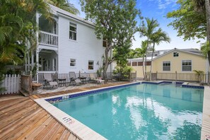 A pool area with a wooden deck, a white building with a balcony, and a yellow building in the background.