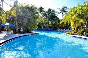 Une piscine à bord incurvé, entourée de palmiers et d’ombrelles bleues.