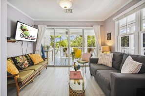 Ein Wohnzimmer mit einer Couch, einem Couchtisch und einem Fernseher. Es gibt einen Balkon mit Blick auf Bäume und einen weißen Zaun.
