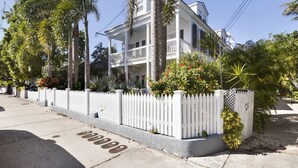 A two-story house with a white picket fence, surrounded by palm trees and various plants. - Lazy Gecko by AvantStay | Walk to Duval St in Key West! | Month Long Stays Only (Key West)