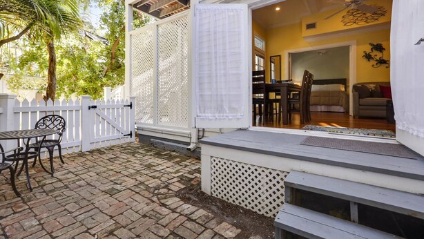 A patio with a table and chairs, a white fence, and a view into a room with a bed and a chair. - Lazy Gecko by AvantStay | Walk to Duval St in Key West! | Month Long Stays Only (Key West)