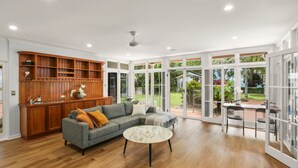 A spacious living room with a grey sectional sofa, a round coffee table, and a wooden built-in shelving unit. Large glass doors lead to a garden view.