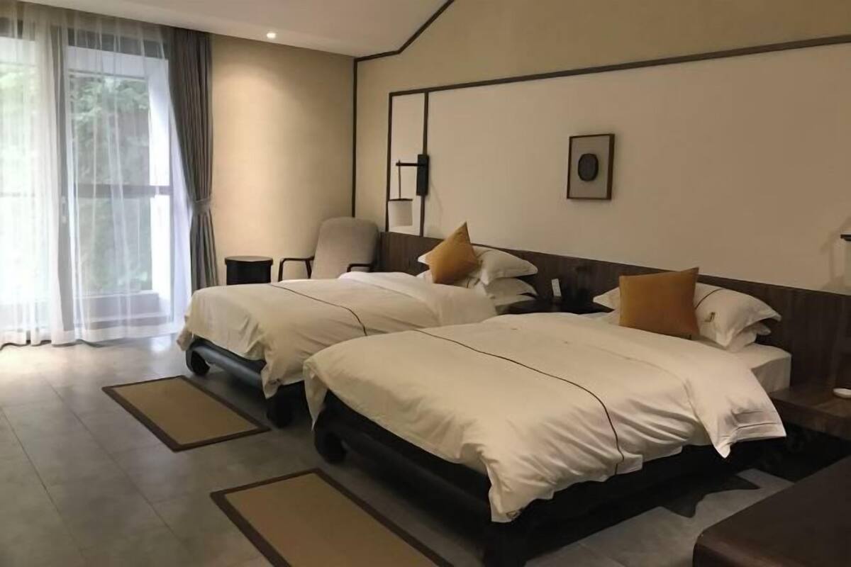 Ett hotellrum med två sängar, en stol, ett skrivbord och ett stort fönster med tunna, genomskinliga gardiner.