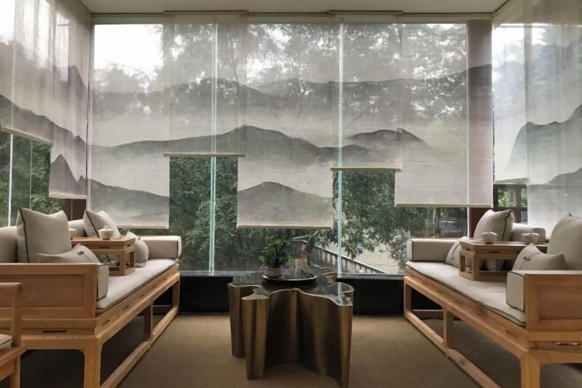 A modern living room with a sofa set, a coffee table, and a view of a mountainous landscape through sheer curtains.