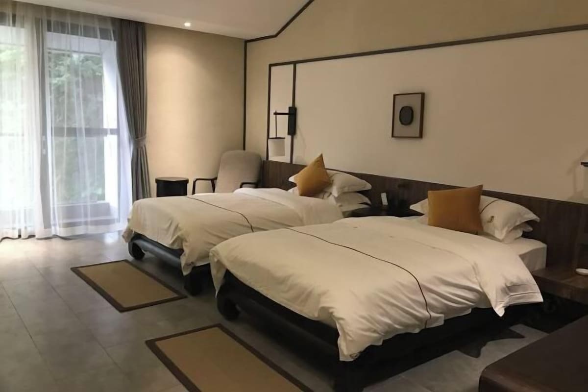 A hotel room with two beds, a chair, a desk, and a large window with sheer curtains.