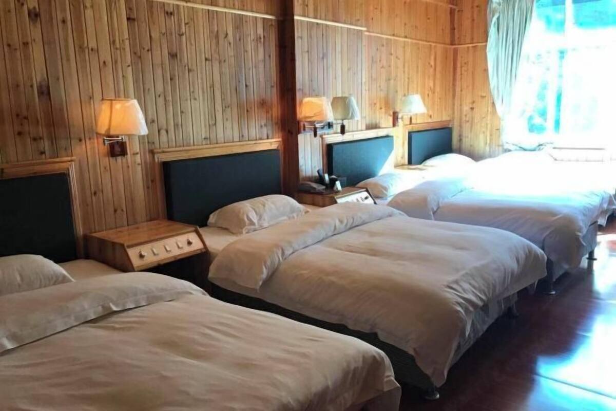 Ett hotellrum med väggar beklädda med träpaneler, tre sängar med vita sängkläder, sänglampor och ett träbord vid sängen med en lampa.