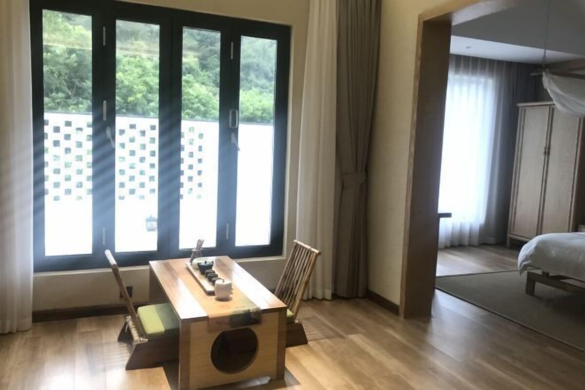 A hotel room with a wooden table, a chair, a bed, and a view of greenery through large windows.