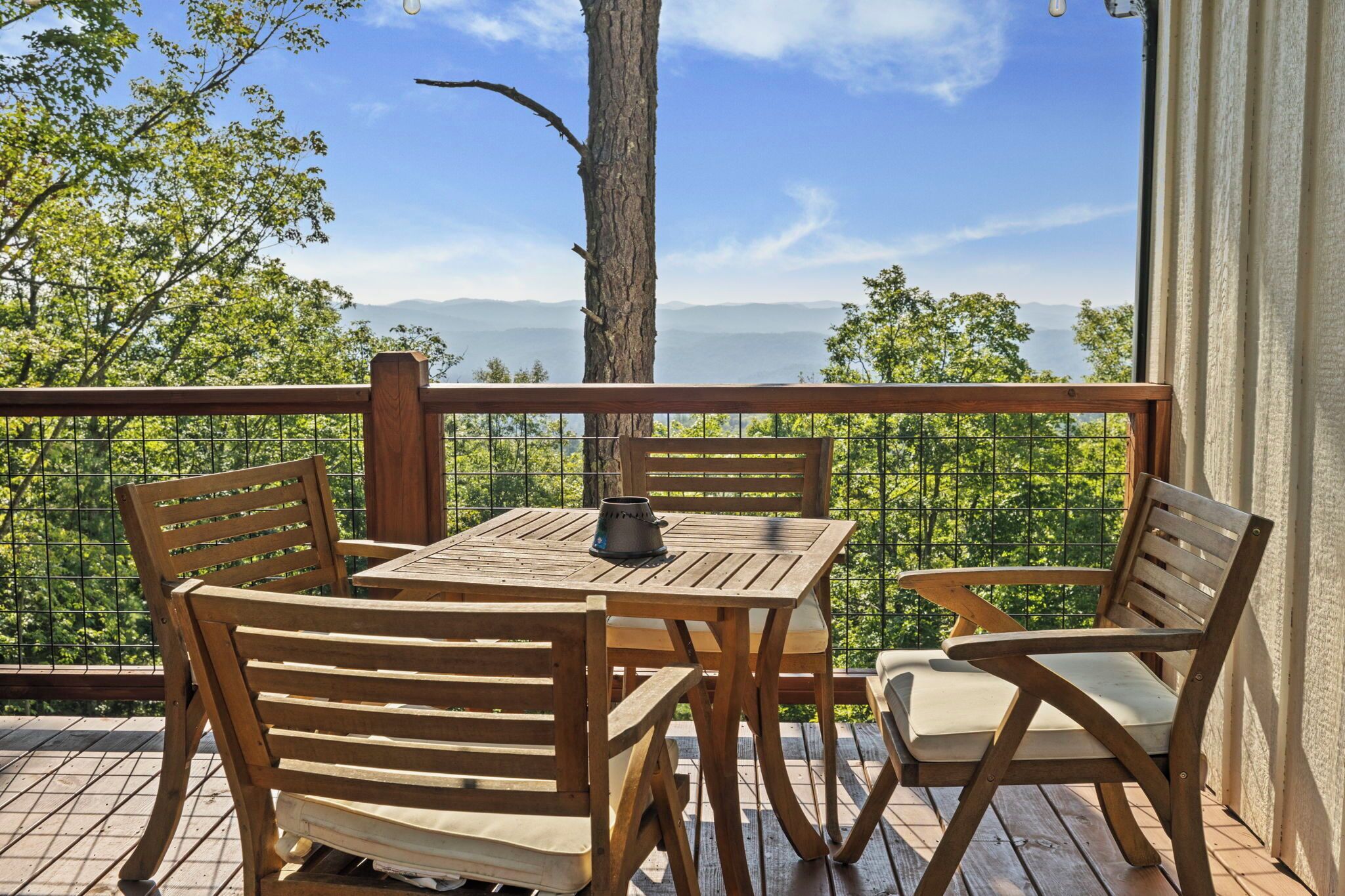 A wooden deck with a table and chairs, overlooking a scenic view of trees and mountains.
