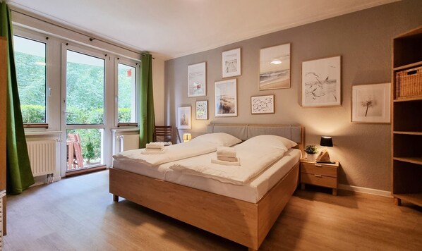 A bedroom with a large bed, a nightstand, a lamp, and a view of greenery through the window.