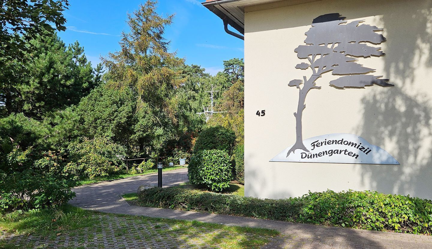 Entrada de un edificio con un letrero en silueta de árbol, sendero pavimentado y abundante vegetación.