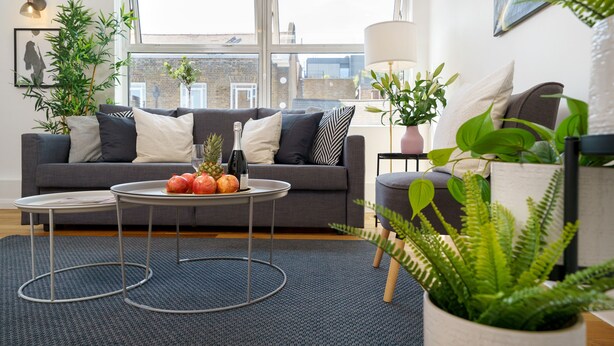 Een moderne woonkamer met een grijze bank, een ronde salontafel met fruit, en een groot raam met uitzicht op gebouwen.