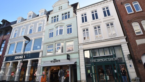 Vista de una calle con una hilera de edificios, incluyendo una tienda llamada 'Schuhka' y 'The Body Shop'.