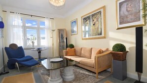 Una sala de estar con un sillón azul, un sofá beige, una mesa de centro de vidrio y un ventanal con cortinas.