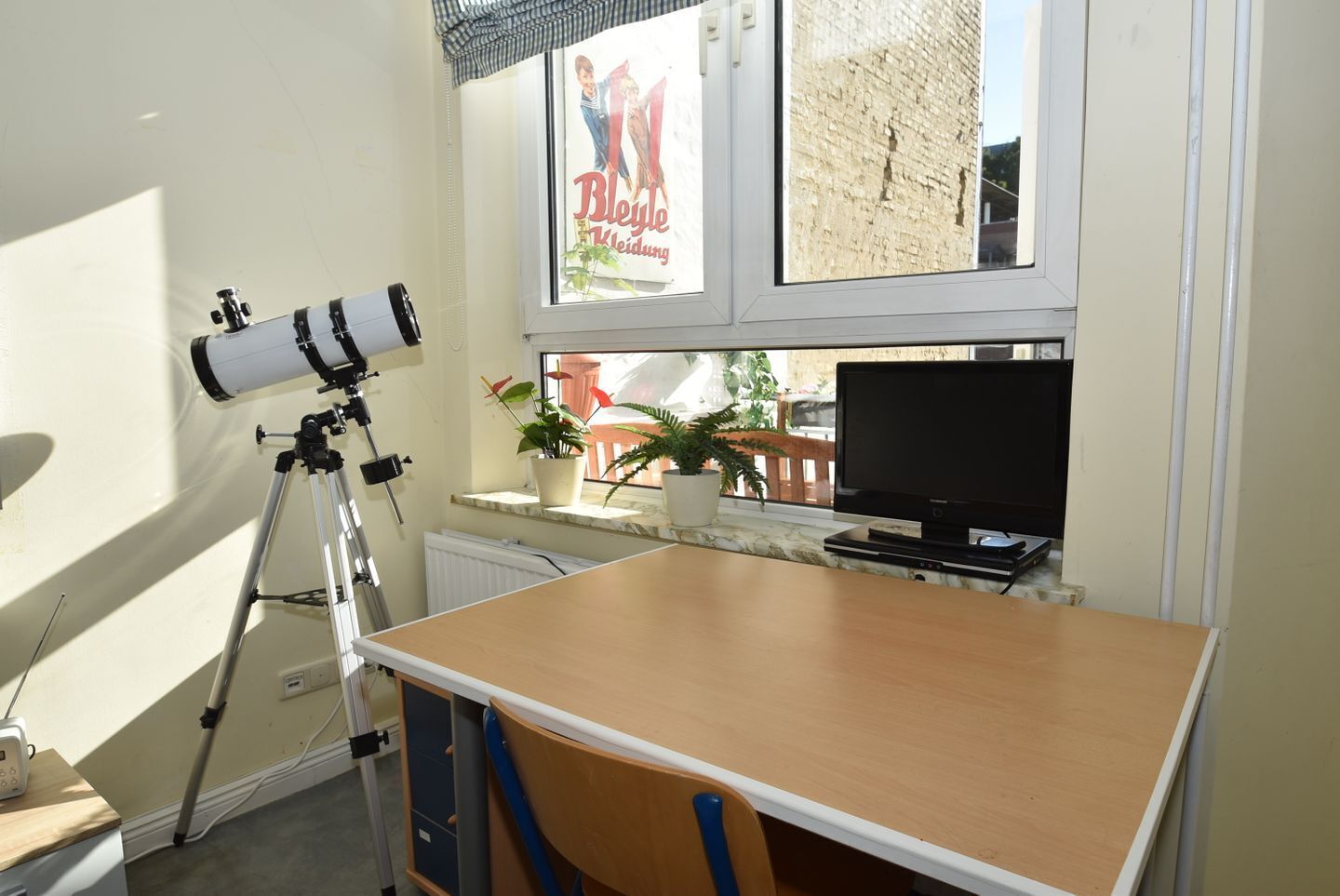 A room with a telescope, a television, a wooden table, and a window with a view of a brick wall and plants.