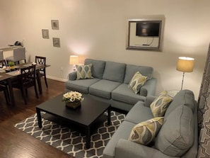 Un salon avec un canapé gris, une table basse agrémentée d’un arrangement floral, une table à manger avec des chaises et un téléviseur fixé au mur.