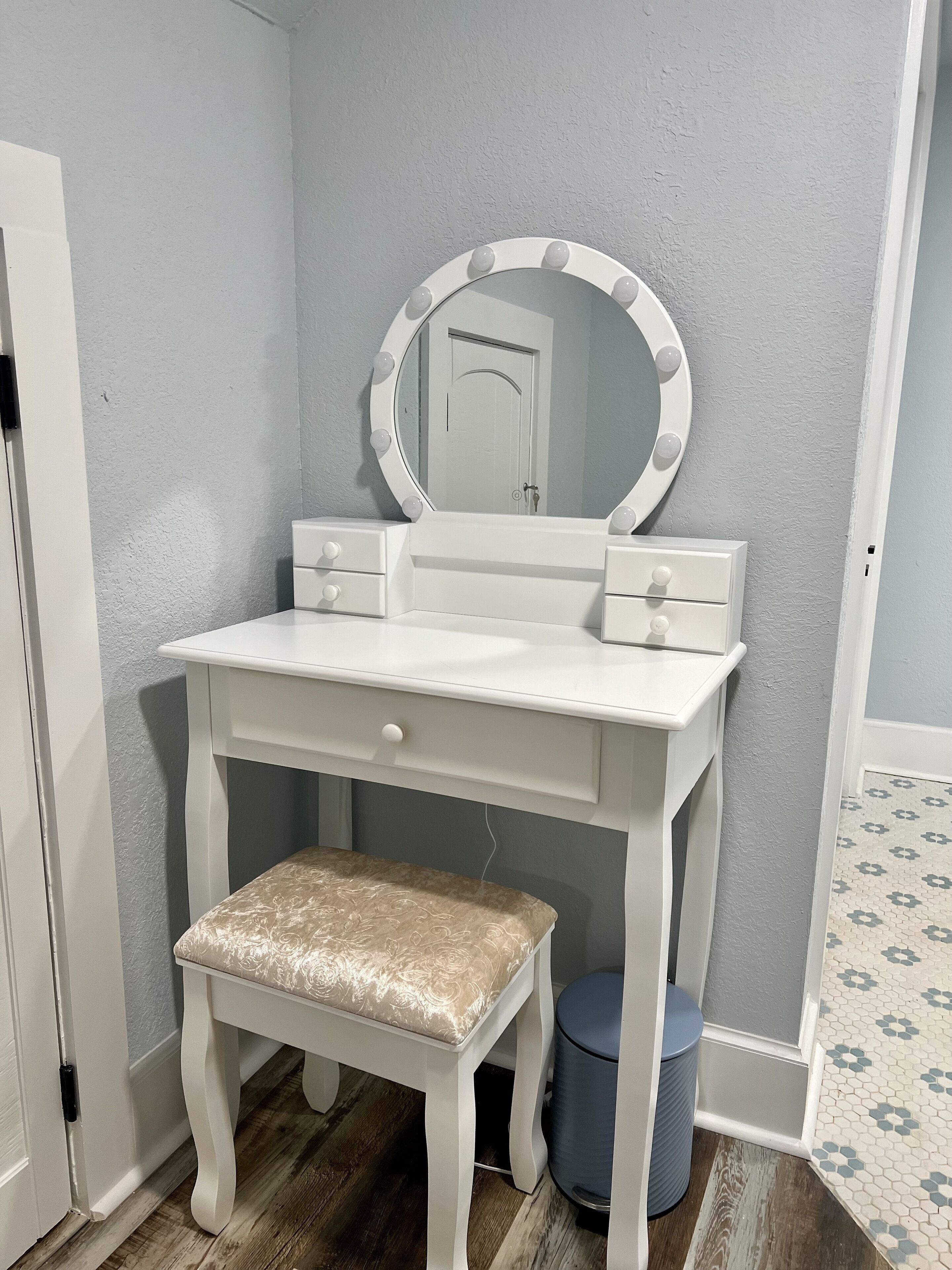 A white vanity with a round mirror and lights, a small bench, and a trash can.