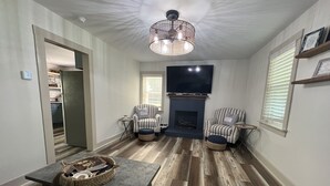 A living room with a fireplace, two striped armchairs, a television, and a dining table with a basket. - Riverside Relaxer Cottage w/Hot Tub+LRG deck+fireplace, w/ Little TN River View (Franklin)