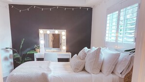 Un dormitorio con cama, tocador con luces, ventilador de techo y ventana con persianas.