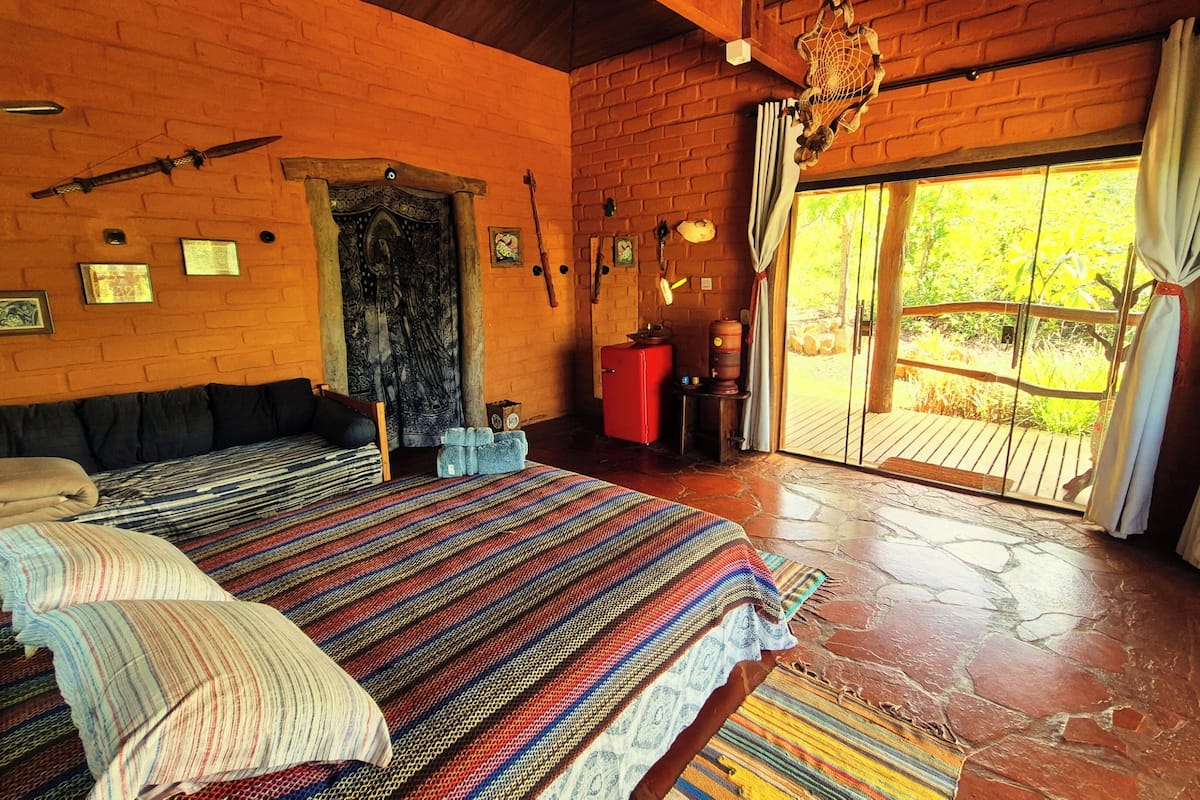 Quarto rústico com uma cama grande, cabeceira de madeira, um armário vermelho e vista para o exterior através de uma porta de vidro.