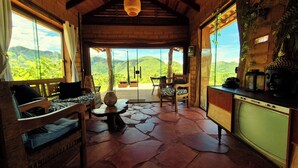 A cozy room with a stone floor, wooden furniture, a large window with a view of green hills, and a Buddha statue on the wall. - Ecopousada Toca do Guará (Alto Paraíso de Goiás)