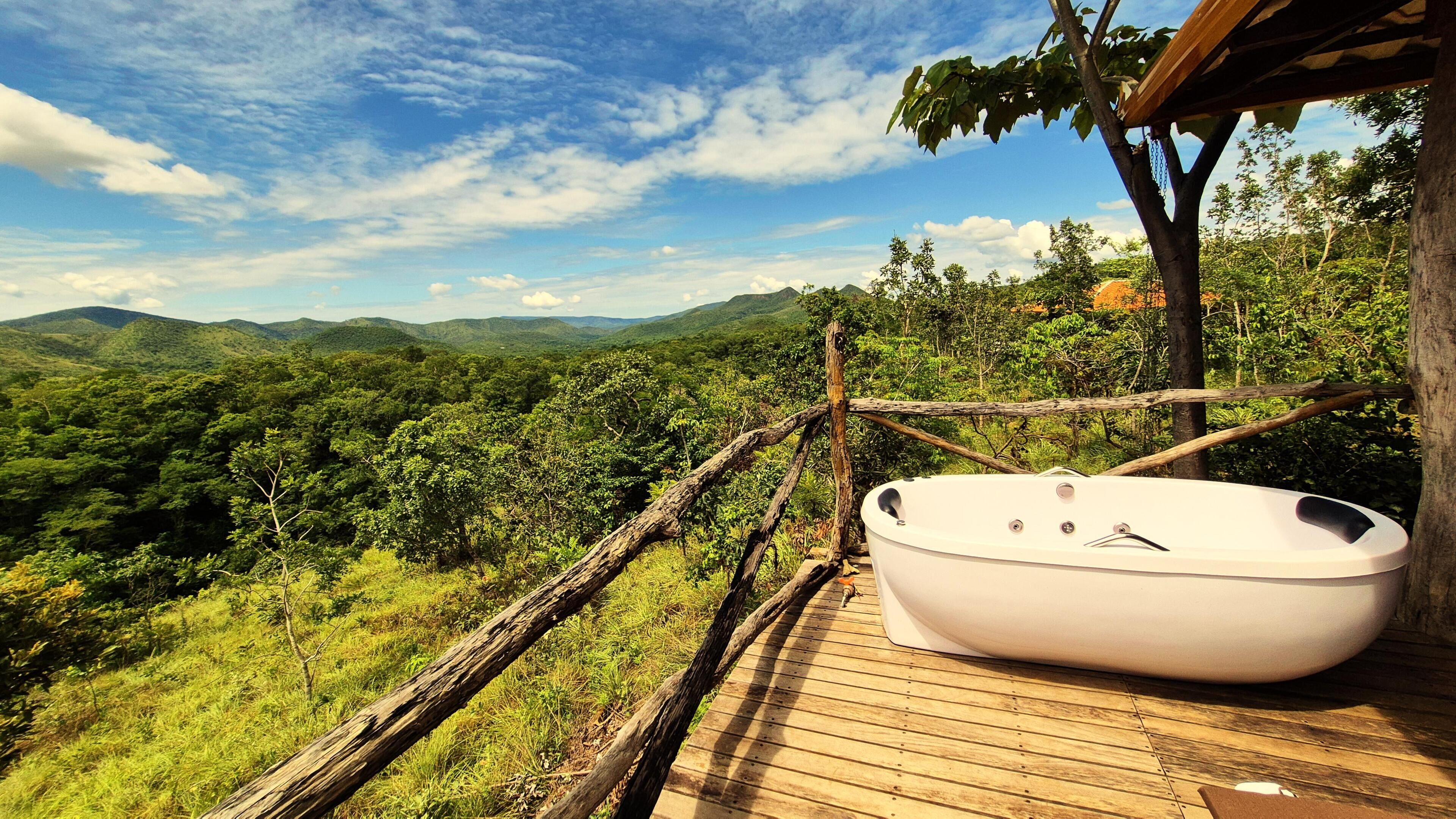 Une terrasse en bois avec une baignoire blanche, entourée d’une végétation luxuriante et dominant un paysage montagneux.