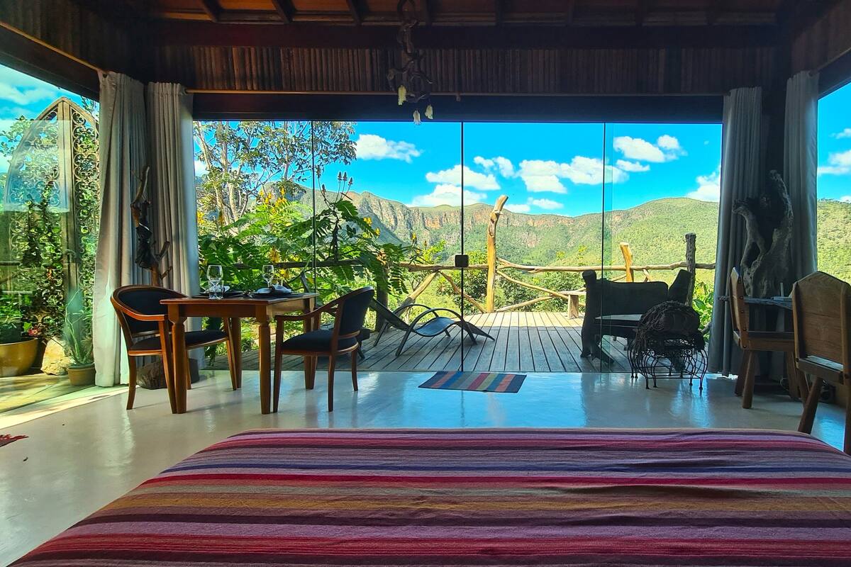 Quarto com uma cama grande, uma mesa de jantar e cadeiras. Vê-se uma vista de montanhas e vegetação através de grandes janelas.