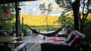 Un sillón hamaca con un cojín colorido, mesas de madera y bancos en una terraza de madera con vistas a un paisaje amarillento con árboles y colinas.