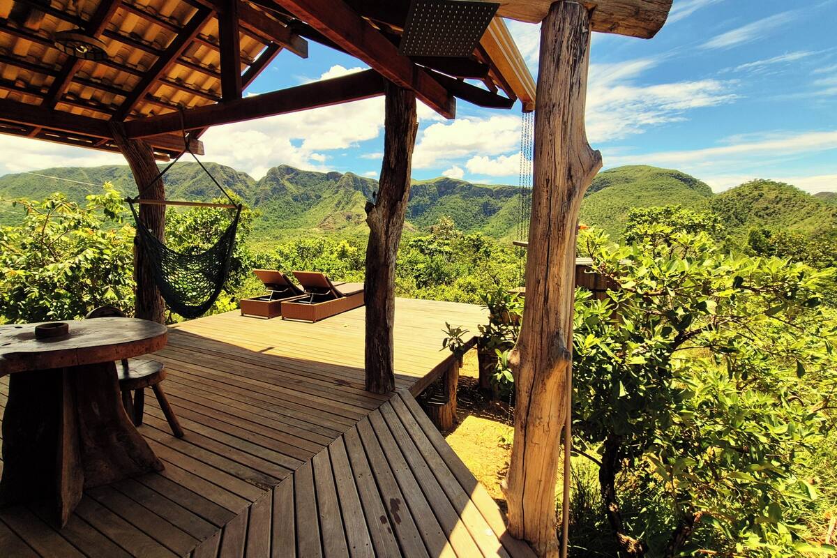 Um deck de madeira com uma mesa e cadeiras, com vista para uma paisagem verde exuberante e montanhas ao longe.