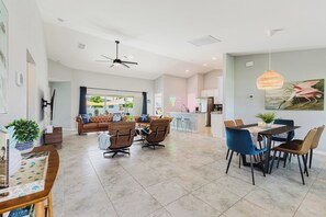 Een ruime woonkamer met een grote bank, een eettafel en stoelen, een keukenhoek en een uitzicht door een glazen deur naar buiten.